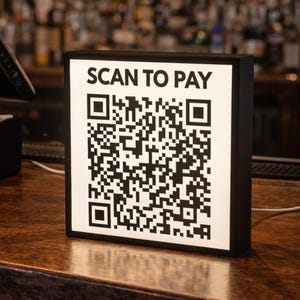 Może przedstawiać: Czarno-biały znak "SCAN TO PAY" z kodem QR jest wyświetlany na drewnianym barze. Czarny system POS znajduje się po lewej stronie. Tło jest rozmyte, pokazując półki z butelkami.