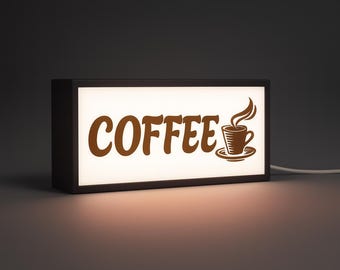 May include: A rectangular, illuminated sign with the word "COFFEE" in brown, stylized lettering. A brown coffee cup with steam is to the right. The sign is in a black frame and has a white interior.