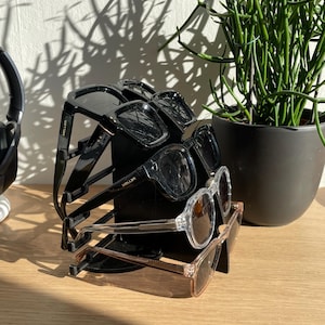 Peut inclure: Une pile de cinq lunettes de soleil sur un support noir. Les lunettes de soleil sont un mélange de montures noires et transparentes. Le support porte l'inscription "Meller".
