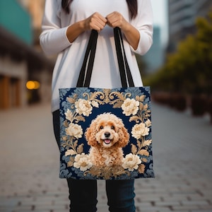 May include: A navy blue tote bag with a smiling, curly-haired dog surrounded by cream-coloured flowers and gold accents. The bag has black handles and a floral pattern background. The dog's fur is light brown.