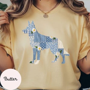 Puede incluir: Camiseta de color mantequilla con un diseño de perro patchwork en tonos azules y blancos. La silueta del perro está compuesta por varios estampados florales y de cuadros, con detalles de margaritas. La palabra "Butter" está impresa en un círculo rosa claro.