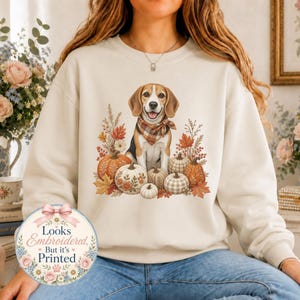 May include: A cream-coloured crewneck sweatshirt featuring a graphic of a beagle wearing a plaid bandana, surrounded by pumpkins and autumn foliage. A circular badge on the lower left displays the text: Looks Embroidered, But it's Printed.