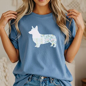 Puede incluir: Camiseta azul con una silueta de corgi blanca rellena de un estampado floral. El estampado floral incluye flores rosas, azules y amarillas. La camiseta es de material suave y tiene mangas cortas.