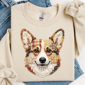 Może przedstawiać: Kremowa bluza z patchworkowym wzorem przedstawiającym pysk psa rasy Corgi. Twarz psa składa się z różnych wzorów tkanin w odcieniach brązu, beżu i bieli. Bluza ma okrągły dekolt i długie rękawy.