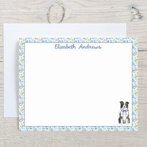 May include: A personalized stationery set featuring a white notepad with a floral border and the name "Elizabeth Andrews" in navy blue script. A watercolor illustration of a border collie with a blue bow tie is in the lower right corner.