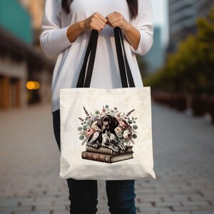 May include: Cream tote bag with black handles. The bag displays a graphic of a dog resting on books, encircled by flowers. The dog has black and white spots and brown eyes.
