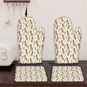 May include: A pair of oven gloves and two matching pot stands. The kitchen textiles display a repeating pattern of German Shepherd dogs, purple flowers, and green foliage on a cream background. Black trim outlines the pot stands.
