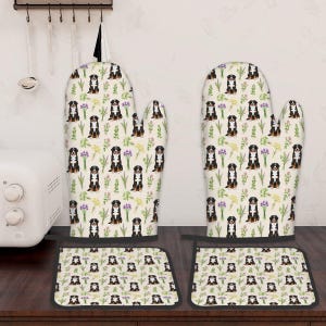 May include: A cream-coloured oven mitt and pot holder set featuring a pattern of Bernese Mountain Dogs, purple and yellow flowers, and green foliage. The set is designed for kitchen use.