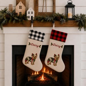 May include: Two off-white Christmas stockings featuring a Yorkshire Terrier illustration, holly accents, and the name Daisy embroidered in red. One stocking has a black and white buffalo check cuff, while the other has a red and black buffalo check cuff.