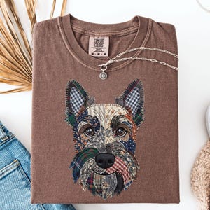 May include: A brown Comfort Colors t-shirt featuring a patchwork Scottish Terrier dog design. The dog's face is made of various fabric patterns in blue, green, red, and white. A silver necklace with a pendant rests on the shirt.