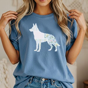 Puede incluir: Camiseta azul con una silueta blanca de perro, estampada con flores y cuadros vichy. El diseño del perro es el elemento principal, sobre un fondo azul liso. La camiseta tiene mangas cortas y cuello redondo.