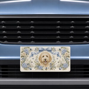 May include: A rectangular license plate frame featuring a central illustration of a golden-colored doodle dog surrounded by blue and white floral arrangements and a light blue ribbon bow, all set against a cream-colored background with four screws.
