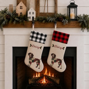 May include: Two cream-colored Christmas stockings featuring a brown and white dog illustration with a colorful patchwork pattern. One stocking has a black and white buffalo check cuff, the other a red and black cuff. Both feature the name Daisy in red.