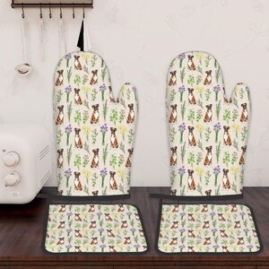 May include: A kitchen set featuring oven gloves and pot holders. The beige fabric is decorated with boxer dog illustrations, purple and yellow flowers, and green leaves. The set has black edging.