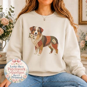 May include: A cream-coloured crewneck sweatshirt featuring a graphic of an Australian Shepherd dog composed of various floral and geometric patchwork patterns in earth tones, accented with white daisy flowers on the neck and hindquarters.