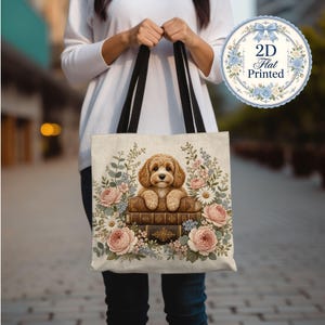 Op de afbeelding: Een beige draagtas met zwarte hengsels voorzien van een gedrukte illustratie van een goudbruine puppy die rust op een stapel van drie oude boeken, omringd door roze rozen, witte madeliefjes en groen gebladerte. Tekst 2D Flat Printed zichtbaar.