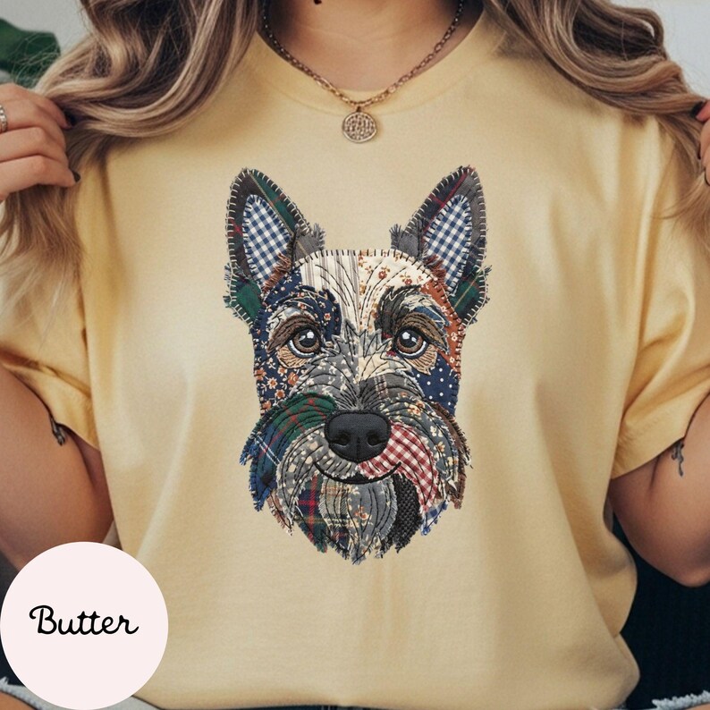 May include: Butter-colored t-shirt featuring a detailed, patchwork-style illustration of a Scottish Terrier dog. The dog's face is composed of various fabric patterns in shades of blue, green, red, and white. The word "Butter" is printed on a light pink circle.