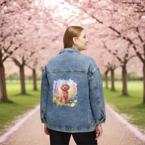 May include: A denim jacket with a colorful illustration of a poodle in a field of flowers. The jacket is a light blue wash with a classic collar and button-up front. The poodle illustration is the focal point.