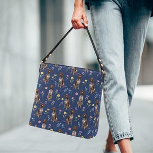May include: A navy blue shoulder bag with a repeating pattern of brown and white dogs and colorful flowers. The bag has a black strap and gold-colored hardware. The bag is held by a person.