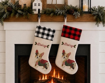 Op de afbeelding: Twee crèmekleurige kerstsokken met een illustratie van een Corgi met een hulsttakje, de naam Daisy in rode schrijfletters en een geruite boord. De ene sok heeft een zwart-witte ruitboord, de andere een rood-zwarte ruitboord.