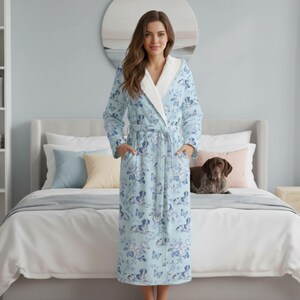 May include: A light blue robe with a floral and dog pattern. The robe has a white trim on the hood and lapels, a matching belt, and pockets. The robe is worn by a person standing in front of a bed.