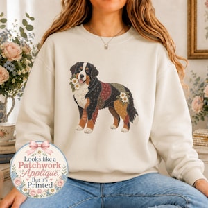 Puede incluir: Una sudadera de cuello redondo color crema con un gráfico de un perro de montaña de Berna con un pelaje estilo patchwork y detalles de margaritas blancas. Una insignia circular abajo a la izquierda muestra el texto: Looks like a Patchwork Applique But it's Printed.