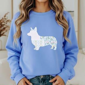 Peut inclure: Un sweat-shirt bleu clair avec un motif de corgi. La silhouette du corgi est remplie de motifs floraux et vichy en blanc, bleu et rose. Le sweat-shirt est de style décontracté. Le design est une illustration charmante et fantaisiste.