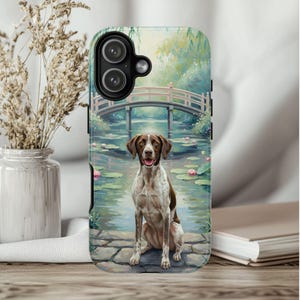 May include: A phone case featuring a painting of a dog sitting by a pond with a bridge in the background. The dog is brown and white, and the scene includes water lilies and a pink bridge.