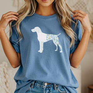 Pode incluir: Camiseta azul clara com uma silhueta de cachorro preenchida com padrões florais e xadrez vichy. O design do cachorro é branco e o padrão floral inclui azul, amarelo e verde. A camiseta é feita de um material macio.