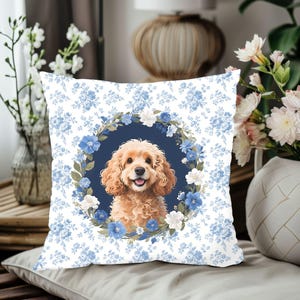 Op de afbeelding: Vierkant kussen met een bloemenpatroon in wit en lichtblauw. Het midden toont een portret van een Cocker Spaniel hond omringd door een krans van blauwe en witte bloemen. Het kussen ligt op een neutraal gekleurd oppervlak.