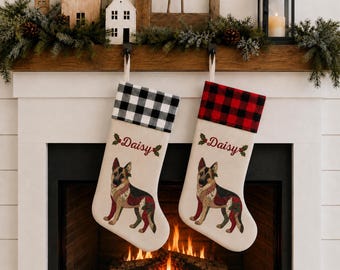 Op de afbeelding: Twee off-white kerstsokken met een illustratie van een Duitse herder, de naam Daisy in rode letters en hulstaccenten. De ene sok heeft een zwart-wit geblokte boord, de andere een rood-zwart geblokte boord.