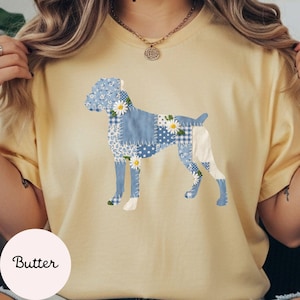 May include: A light yellow t-shirt featuring a patchwork dog design in shades of blue and white, adorned with daisy accents. The word "Butter" is printed in a circular design at the bottom left.