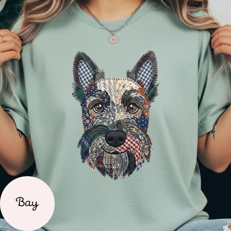 May include: A light green t-shirt featuring a patchwork design of a Scottish Terrier dog. The dog's face is made up of various fabric patterns, including plaid and floral prints. The word "Bay" is printed on a white circle.
