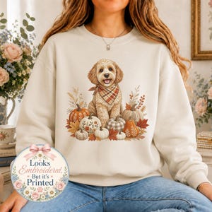 May include: A cream-colored crewneck sweatshirt featuring a graphic of a golden-colored dog wearing a plaid bandana, surrounded by various pumpkins and autumn leaves. A circular badge on the lower left reads, Looks Embroidered, But it's Printed.