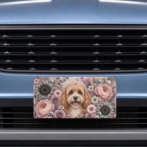 May include: A rectangular license plate frame featuring a central illustration of a light brown dog with floppy ears surrounded by a floral pattern of pink peonies, white daisies, and dark gray sunflowers, mounted on a blue vehicle grille.