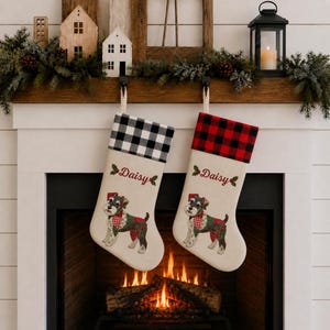 May include: Two off-white fabric Christmas stockings featuring the name Daisy in red script, accented by holly leaves. One stocking has a black and white buffalo check cuff, while the other has a red and black buffalo check cuff, both with dog graphics.