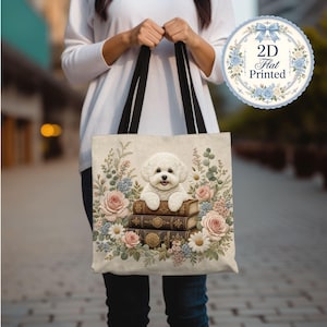 May include: A beige tote bag with black handles featuring a printed illustration of a white Bichon Frise dog resting on a stack of three vintage-style books, surrounded by a colorful arrangement of pink roses, white daisies, and green foliage.