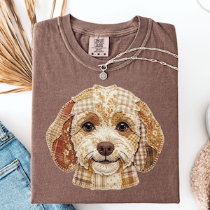 May include: A brown Comfort Colors t-shirt featuring a patchwork dog face design. The dog's face is made of various patterned fabrics in shades of brown, cream, and orange. A silver necklace with a pendant is draped across the shirt.