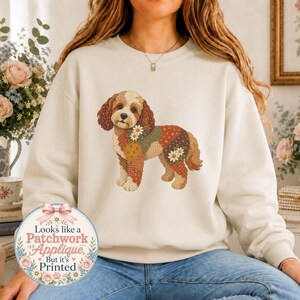 Puede incluir: Una sudadera de cuello redondo color crema con un gráfico central de un perro con un pelaje estilo patchwork en tonos marrón, naranja y verde, decorado con flores blancas. Una insignia circular en la parte inferior izquierda dice: Looks like a Patchwork Applique, But it's Printed.