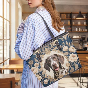 May include: A blue tote bag with a German Shorthaired Pointer portrait. The bag features a floral and scroll design with gold accents and brown leather straps. The dog has brown and white markings.