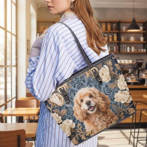May include: A tote bag with a floral and dog design. The bag features a smiling Cocker Spaniel on a blue background with cream-colored flowers and gold accents. It has a black strap and gold buckles.