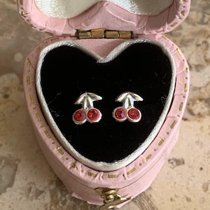 Könnte beinhalten: Ein Paar silberne Kirsch-Ohrringe mit roten Kristall-Akzenten, präsentiert in einer rosafarbenen herzförmigen Schachtel.