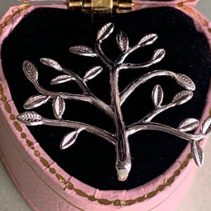 Könnte beinhalten: Silberfarbene baumförmige Brosche mit detaillierten Blättern, die in einer rosa herzförmigen Schmuckschatulle mit schwarzem Innenraum liegt. Die Schatulle hat goldfarbene Verzierungen und einen goldenen Verschluss.