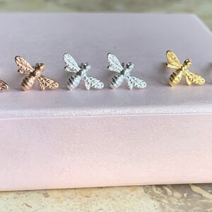 Può includere: Sei orecchini a forma di ape in una scatola rosa. Due sono d'oro, due d'argento e due d'oro rosa.