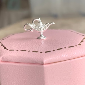 Könnte beinhalten: Ein silberner Dschinnenlampen-Anhänger in einer rosa Geschenkbox mit goldenen Verzierungen.