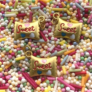 Könnte beinhalten: Drei goldfarbene Charms in Form von Schokoriegeln mit dem Wort "Sweet" darauf. Die Charms sind von bunten Streuseln umgeben.