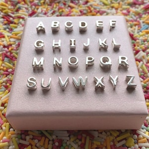Könnte beinhalten: Ein Satz silberner Buchstaben-Anhänger in einer rosa Box. Die Buchstaben sind in alphabetischer Reihenfolge von A bis Z angeordnet.