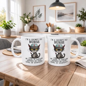 Op de afbeelding: Twee witte keramische mokken met een afbeelding van een norse grijze kat met een verpleegsterskapje die een injectiespuit vasthoudt. De tekst luidt: I'M A GRUMPY OLD NURSE. MY LEVRL OF SARCASM DEPENDS ON YOUR LEVEL OF STUPIDITY. Mokken met een oor.