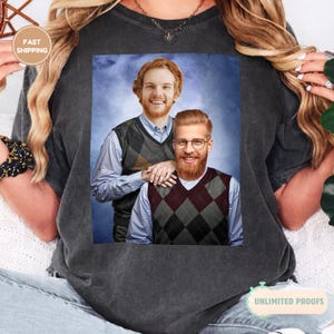 May include: Dark gray t-shirt featuring a portrait of two men with red hair, wearing argyle vests over light blue shirts. One man wears glasses and has a beard. The background is a blue sky. The shirt has the words "UNLIMITED PROOFS".