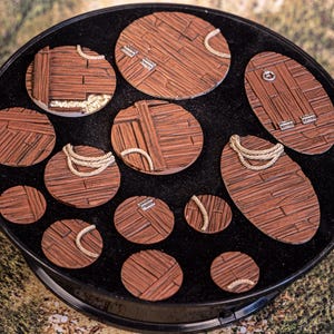 Könnte beinhalten: Eine Sammlung von zwölf runden und ovalen Miniaturbasen mit einer braunen Holzplankenstruktur. Einige Basen enthalten kleine silberne Metallscharniere oder beigefarbene Seilschlaufen. Das Set ist in einem schwarzen runden Tablett angeordnet.